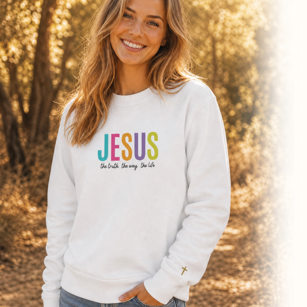 A woman smiling outdoors in a white crewneck sweatshirt with a colorful 'JESUS' graphic and the phrase 'Who Sings, Saves Us' printed on the chest, paired with blue jeans.
