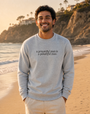 A smiling man standing on a sandy beach at sunset wearing a light gray crewneck sweatshirt with the black printed text 'a prayerful man is a powerful man' on the chest, paired with light beige pants.