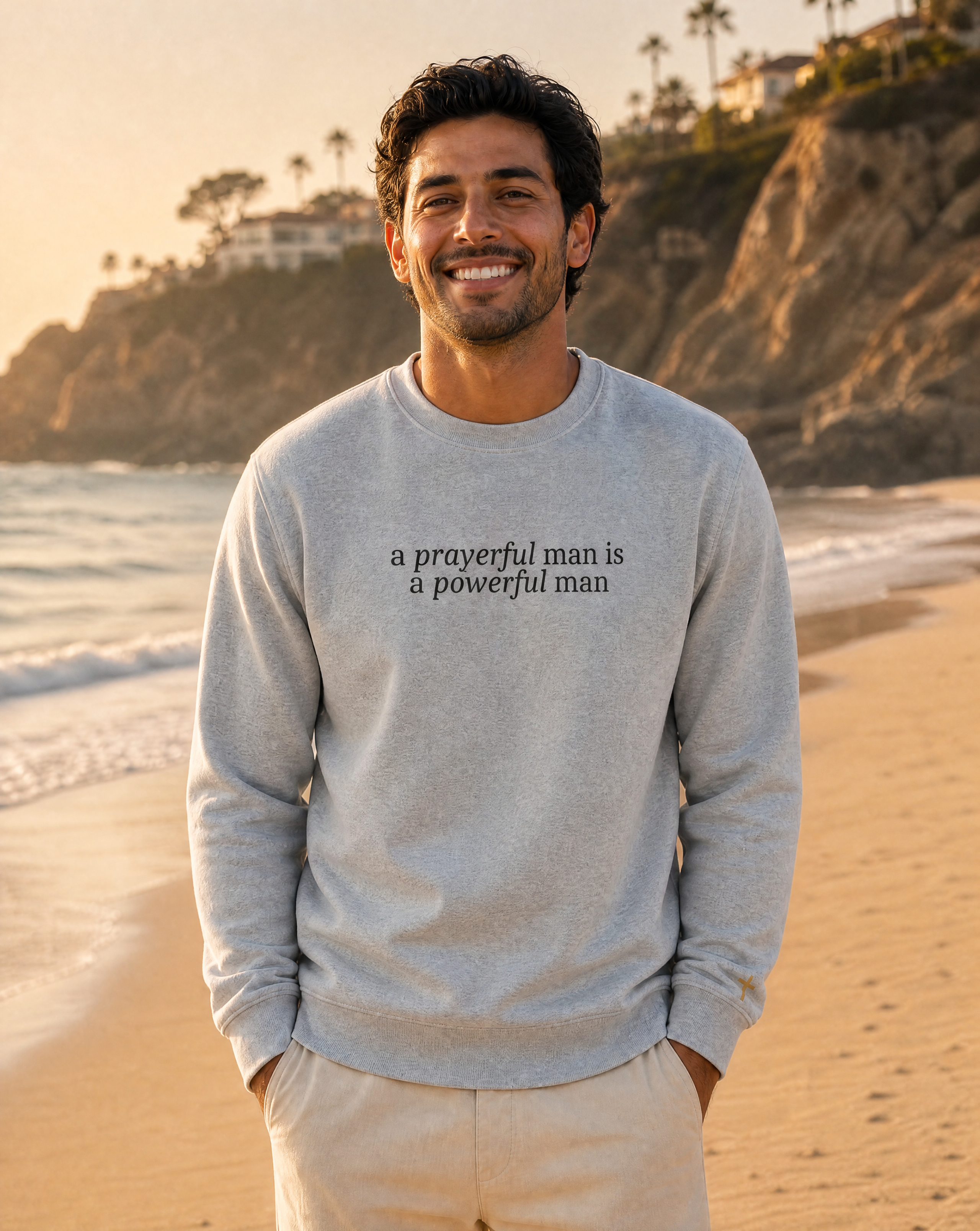 A smiling man standing on a sandy beach at sunset wearing a light gray crewneck sweatshirt with the black printed text 'a prayerful man is a powerful man' on the chest, paired with light beige pants.